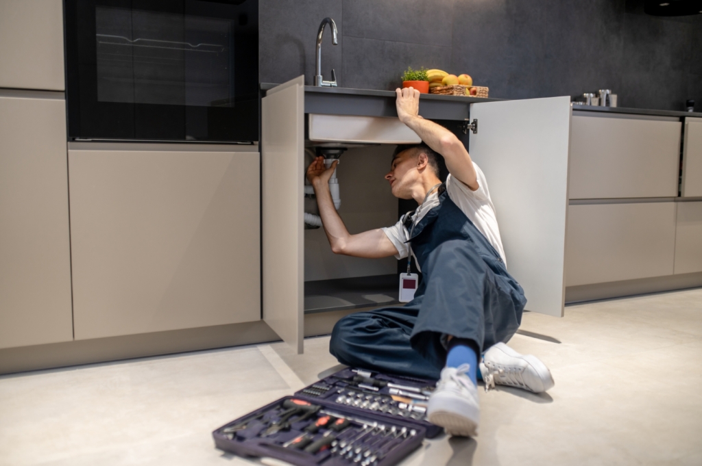 Haushaltsreparatur-Mann mittleren Alters-inspiziert-Rohre-berühren-handspüle-stilvolle-moderne-küche